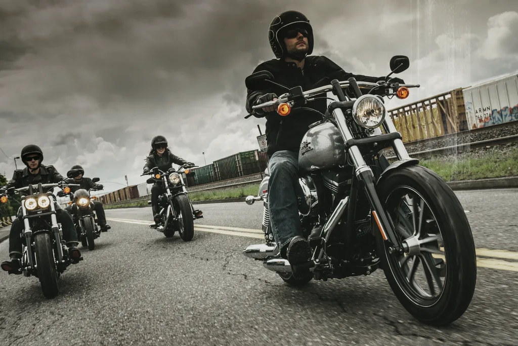 A rider on a silver Harley-Davidson Dyna Street Bob at the front of a group of four motorcycles on a road beside railway wagons.