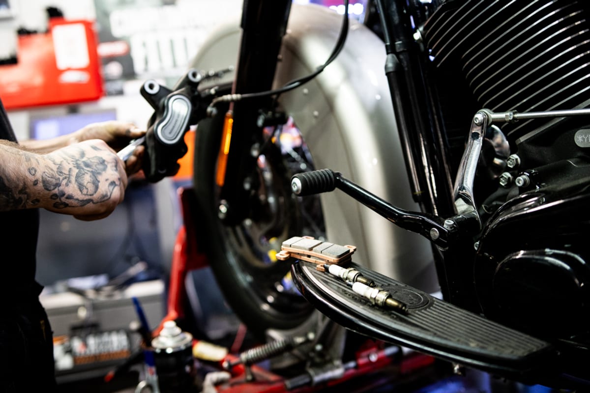 A mechanic inspecting a Harley-Davidson brake caliper with a fresh set of pads ready on the floorboard